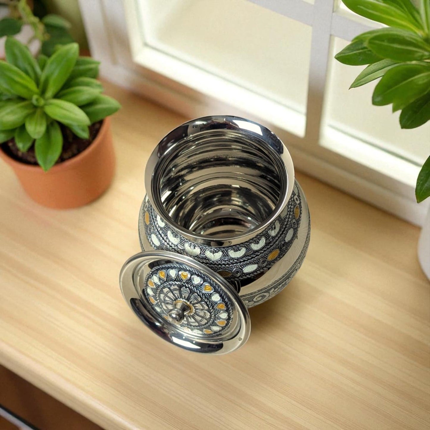 Antique Stainless Steel Lota with intricate design and lid placed on wooden surface near green plants