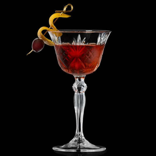 RCR Melodia Coupette Glass filled with a cocktail garnished with orange twist and cherry, on a dark background