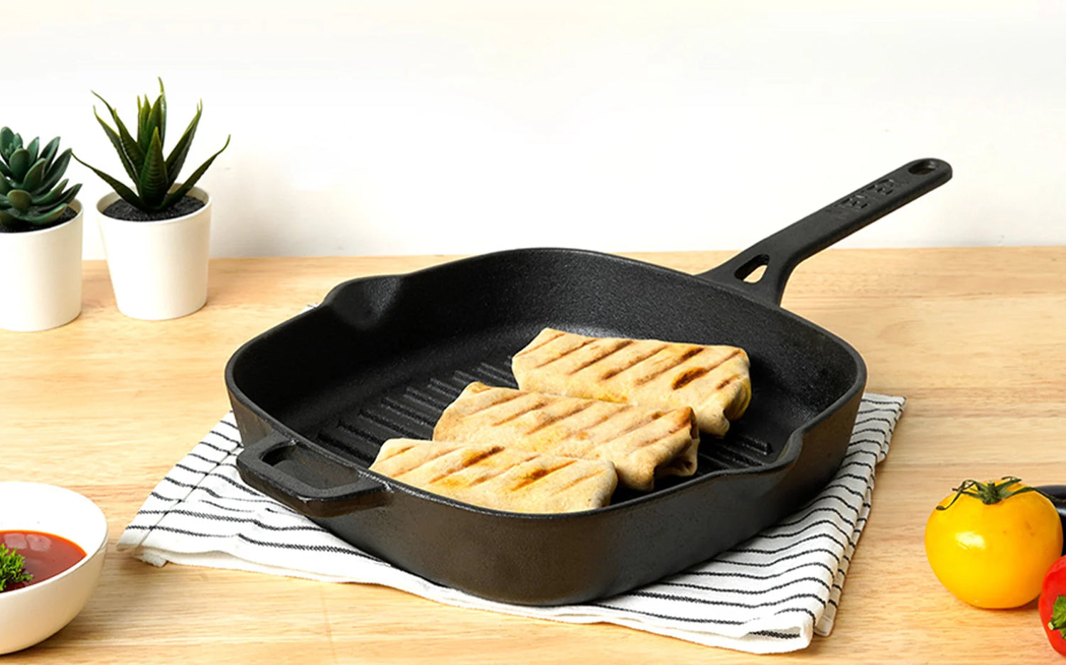 Meyer cast iron grill pan 25cm with grilled food on a wooden table with plants and a white wall in the background.