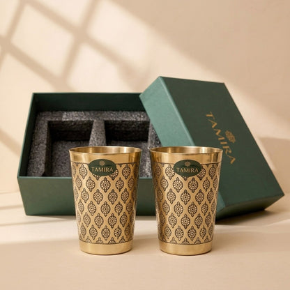 Two pure brass glasses with a black floral motif in front of their green gift box